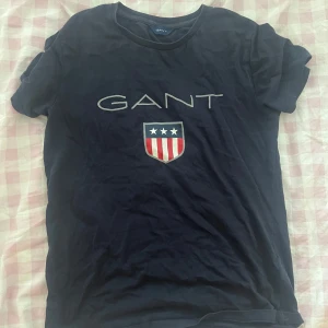 Mörkblå GANT t-shirt med tryck - Snygg mörkblå t-shirt från GANT med stort logotryck och sköld på bröstet. Klassisk rund halsringning och korta ärmar. Tillverkad i mjuk bomull som är skön att bära hela dagen. Perfekt för en clean och sportig look.
