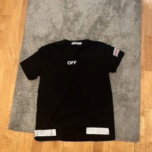 Svart Off-White t-shirt med tryck - Svart t-shirt från Off-White med vit text 'OFF' på bröstet och stort ikoniskt grafiskt tryck på ryggen. T-shirten har korta ärmar, rund hals och vita detaljer nertill. Perfekt för dig som gillar streetwear och vill sticka ut. Passar folk mellan 172-183 cm.