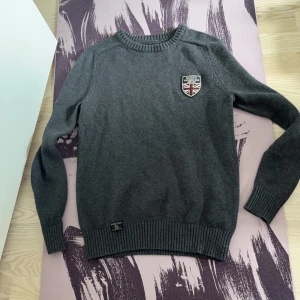 Grå stickad tröja från Morris Gents - Mörkgrå stickad tröja från Morris Gents med rund halsringning och ribbade muddar. Tröjan har ett snyggt broderat märke med brittisk flagga på bröstet och en diskret etikett vid nederkanten. Perfekt för dig som gillar klassisk och stilren look.