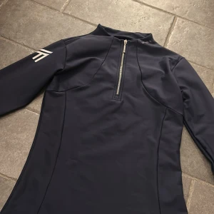 Marinblå half zip tröja från Maximilian equestrian - Sportig marinblå långärmad tröja från Maximilian Equestrian med half zip framtill och logga på bröstet, ryggen och ärmen. Tröjan är figursydd och tillverkad i ett stretchigt syntetmaterial.