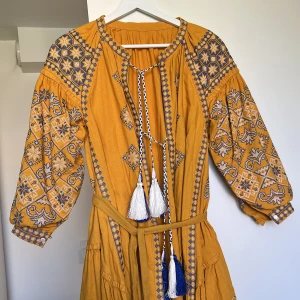 Broderad gul klänning med knytband - Superfin gul klänning med blå och vita broderier, vida trekvartsärmar och knytband med tofsar. Klänningen har bälte i midjan och vackra detaljer över hela plagget. Perfekt för dig som gillar boho-vibe och unika mönster.