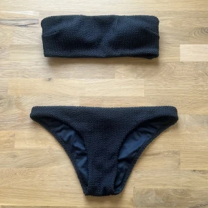 Svart bikini i textur - Svart bikini med textur och enkel design. Överdelen är axelbandslös med metallspänne bak och underdelen har klassisk passform. Båda delarna är storlek 34