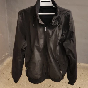 Svart vindjacka från Dressmann L - Svart vindjacka från Dressmann i storlek L. Jackan har dragkedja framtill, hög krage med snörning och ribbade muddar. Tillverkad i polyester, perfekt för blåsiga dagar. Enkel och stilren design som funkar till det mesta.