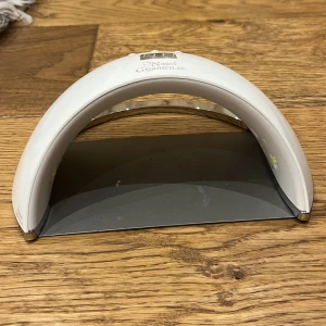 Nagelgiganten UV LED-lampa för naglar - UV LED-lampa från Nagelgiganten i vit plast med digital display och tre knappar för timer (30s, 60s, 99s low heat mode). Bågeformad design med många LED-lampor på insidan och medföljande svart strömadapter. Perfekt för att härda gelnaglar hemma.