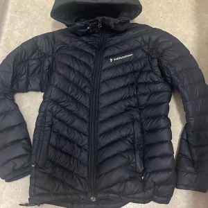 Svart dunjacka Peak Performance S - Säljer en svart dunjacka från Peak Performance i storlek S. Jackan har huva, dragkedja framtill och två fickor med dragkedja. Snyggt mönstrad med chevron-sömmar och logga på bröstet. Perfekt för kalla dagar och riktigt skön att bära.