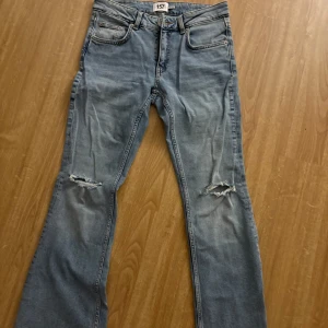 Ljusblå bootcut jeans med slitningar - Säljer ett par ljusblå jeans med bootcut-modell och slitna detaljer på båda knäna. Klassisk femficksdesign och normal passform. Perfekta för en avslappnad och trendig look.