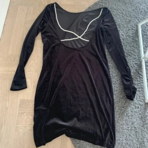 Svart sammetsklänning med pärldetalj - Svart kort sammetsklänning med lång ärm och snygg meshdetalj framtill. Urringningen är dekorerad med pärlor i ett vågigt mönster som ger en unik look. Klänningen har en figurnära passform och känns både lyxig och trendig. Baksidan är med pärlor och öppen rygg (bild nr.1). Använd en gång på nyår.