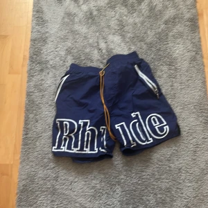 Blå Rhude shorts med snörning - Snygga marinblå shorts från Rhude med stora vita logobokstäver framtill. De har gul snörning i midjan, dragkedjefickor och meshfoder på insidan. Perfekta för sommaren och chill dagar. Stilen är loose och materialet känns syntetiskt och snabbtorkande.