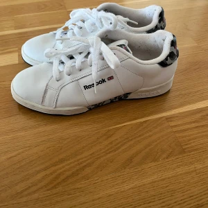 Vita Reebok sneakers med camo-detaljer - Snygga vita Reebok sneakers med svarta och grå camo-detaljer på häl och plös. Klassisk låg modell med rund tå, snörning och platt sula. Skorna är i syntetmaterial och har Reebok-logga på sidan och plösen. Perfekta för en clean och sportig look.