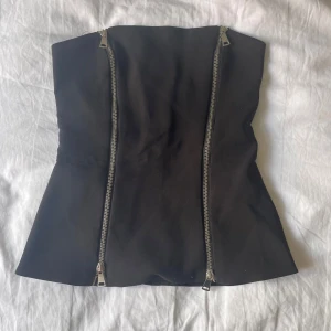 Svart (tube) korsett-topp från Zara XS - Supersnygg svart (tube) korsett-topp från Zara i storlek XS. Toppen är axelbandslös och har två silvriga dragkedjor framtill som ger en edgy vibe. Figurnära passform och lätt utsvängd nertill. Perfekt att styla med jeans eller kjol för en cool look.