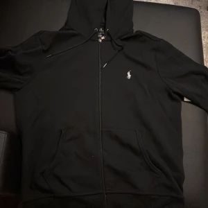 Svart hoodiekofta från Polo Ralph Lauren - Svart hoodiejacka från Polo Ralph Lauren med dragkedja framtill, huva med snörning och klassisk logga broderad på bröstet. Jackan har kängurufickor och är tillverkad i mjukt bomullsmaterial. Perfekt för en chill och stilren look.