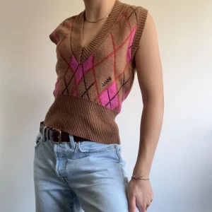 Brun och rosa rutig väst från Ganni - Stickad väst från Ganni i brunt med rutigt mönster. Djup v-ringning och bred ribbad midja ger en snygg passform. Perfekt att styla över en skjorta eller t-shirt för en cool look.