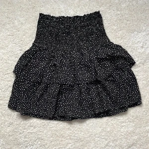 Prickig volangkjol - Supersöt svart kortkjol från Kappahl med vita prickar och volanger. Kjolen har smock i midjan för en bekväm passform och är gjord i ett lätt, flowy material. Storlek xs-s