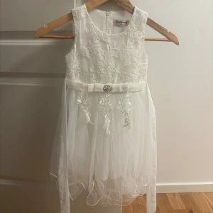 Vit blommig klänning Pretty Baby - Supersöt vit kortklänning från Pretty Baby med broderade blommor, markerad midja med rosett och glittrig dekoration framtill. Klänningen är ärmlös och har en lätt tyllkjol som ger extra volym. Perfekt för speciella tillfällen!