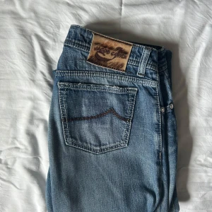 Jacob Cohen Slim Fit Jeans - Snygga blå jeans från Jacob Cohen i storlek 34 men sitter som W31-32 med klassisk femficksdesign och ljus tvätt. Jeansen har en skinnpatch med logga bak i midjan och kontrastsömmar på bakfickorna. Modellen är slimfit och passar perfekt till en avslappnad stil.