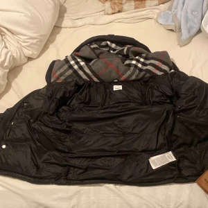 Svart pufferjacka från Burberry - Säljer en svart pufferjacka från Burberry med klassisk logga på ärmen och rutigt foder i grått, vitt och rött. Jackan har hög krage, dragkedja och knappar framtill. Perfekt för kalla dagar och riktigt snygg till streetwear-stilen.
