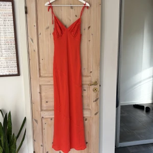 långklänning från Mooloola - Säljer denna super snygga röd/ orangea  långklänning från Mooloola med smala axelband som knyts och v-ringad byst med rynkade detaljer. Klänningen har en figurnära passform och är tillverkad i ett lätt, mjukt tyg som faller fint. Perfekt för sommarens alla tillfällen. Storleken (Australiens storlek) motsvarar ungefär S i Sverige, klänningen är även väligt lång 