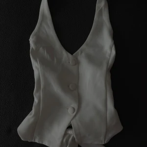 Vit omlottväst med knyt och knappar - Snygg vit väst i omlottmodell med djup v-ringning och tre klädda knappar framtill. Västen har breda axelband och ett brett knytband i ryggen som ger en feminin siluett. Perfekt att styla över en skjorta eller t-shirt för en trendig look.