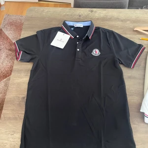Svart Moncler pikétröja  - Svart pikétröja från Moncler med klassisk krage och knäppning framtill. Tröjan har Moncler-logga på bröstet och snygga rödvita ränder på krage och ärmslut. Tillverkad i bomull för en skön känsla. Perfekt för en clean och stilren look.