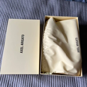 Svarta sneakers från Axel Arigato - Stilrena svarta sneakers från Axel Arigato i mocka med diskret guldtext på sidan. Skorna har en platt sula och elastiskt band över vristen istället för klassisk snörning, vilket ger en clean och modern look. Perfekta för dig som gillar minimalistisk stil.