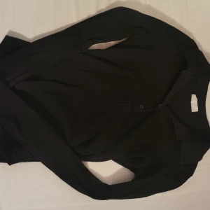 Svart ribbad långärmad pikétröja - Svart långärmad pikétröja med ribbad struktur och klassisk krage. Tröjan har knappar framtill och är perfekt för en stilren och enkel look. Passar dig som gillar minimalistiska plagg med en clean vibe.