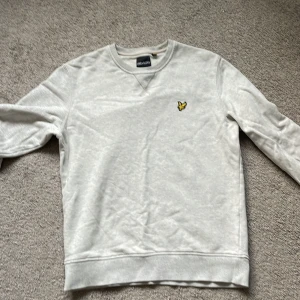 Ljus grå sweatshirt från Lyle & Scott - Vit sweatshirt från Lyle & Scott med klassisk rund hals och broderad gul logga på bröstet. Tröjan har långa ärmar och ribbade muddar vid ärmslut och nederkant. Tillverkad i mjuk bomull för en skön och avslappnad stil.