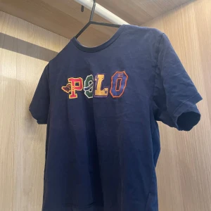 Blå POLO t-shirt från Ralph Lauren - Snygg mörkblå t-shirt från POLO Ralph Lauren med färgglad broderad logga på bröstet. Klassisk passform med rund hals och korta ärmar samt lite cropped. Tillverkad i mjuk bomull som känns skön mot huden.  Passar xs och s