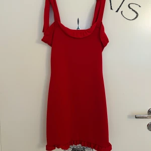 Röd klänning med volangdetaljer från Boohoo - Röd kort klänning med breda axelband och volangdetaljer vid halsringning och nederkant från Boohoo i storlek 34. Axelbanden går att knyta som man vill. Aldrig använd.