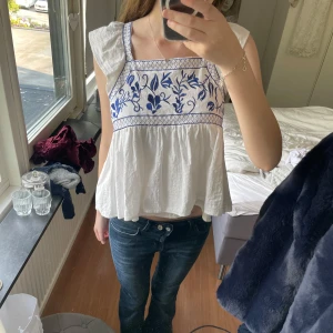 Vit blus med blå broderi och knyt - Supersöt vit blus med blått broderat blommönster upptill och volangig nederdel. Toppen har breda axelband, lätt rynkade ärmar och knyt i ryggen för extra detalj. Perfekt för varma dagar och ger en bohemisk vibe.💞
