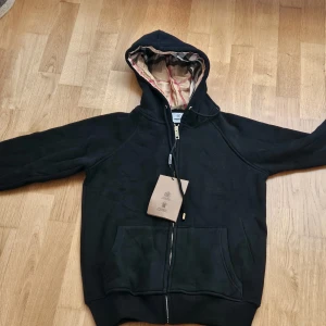 Svart hoodiejacka från Burberry - Pris kan diskuteras kan lösa dunder priser. Svart hoodiejacka från Burberry med klassiskt rutigt foder i huvan. Jackan har dragkedja framtill, snörning i huvan och två stora fickor. Perfekt för dig som vill ha en stilren och lyxig streetlook med ikoniska Burberry-detaljer.