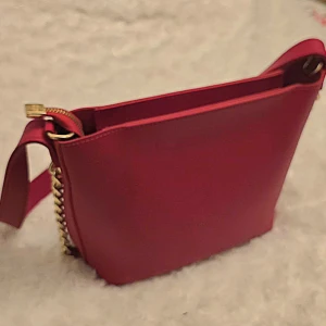 Röd axelväska med guldkedja - Snygg röd axelväska i slät skinnimitation med bred axelrem och guldfärgade kedjedetaljer på sidorna. Väskan har dragkedja upptill och en stilren, enkel design som passar perfekt till din outfit.
