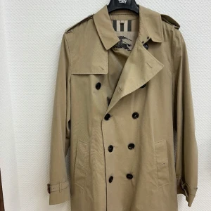 Burberry Men’s Heritage Kensington Short Trench Coat  - Storlek: 52 EU (motsvarar Medium–Large) 	•	Färg: beige 	•	Modell: Kensington – kort längd 	•	Tillverkad i England 	•	Dubbelknäppning, axelklaffar, stormflik, justerbara ärmslut, bälte med metallspännen (D-ringar) och klassiskt rutigt Burberry-foder 	•	Skick: Mycket fint begagnat skick, sparsamt använd och väl omhändertagen 	•	Alla originaldetaljer finns kvar  Nypris ca 23 500 kr