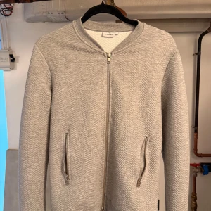 Grå cardigan/kofta från J.Lindeberg - Snygg ljusgrå cardigan kofta från J.Lindeberg i bomullsmix med subtilt mönster.  Perfekt för dig som gillar stilren och enkel design! Skicka gärna ett dm ifall du vill ha fler bilder!