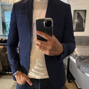 Blå rutig kavaj från Isaac Dewhirst - Snygg marinblå kavaj från Isaac Dewhirst med diskret rutmönster. Klassisk passform med två knappar framtill, fickor med lock och innerfoder med mönster. Perfekt för dig som vill ha en stilren och modern look.