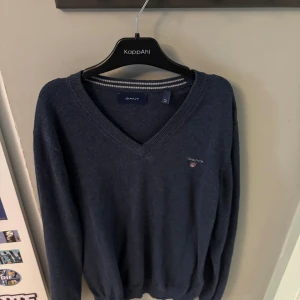 Mörkblå v-ringad tröja från Gant - Klassisk mörkblå v-ringad tröja från Gant med broderad logga på bröstet. Tröjan har långa ärmar och ribbade muddar vid ärmslut och nederkant. Perfekt för en stilren och avslappnad look.