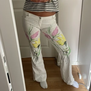 Vita blommiga jeans från Estensivo - Unika vita jeans från Estensivo med blommönster i gult, rosa och grönt framtill. Byxorna har raka ben, låg midja och coola små nitar vid fickorna. Perfekt för dig som vill sticka ut med en färgglad och kreativ look.