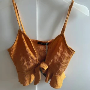 Senapsgul croppad linnetopp med knyt - Superfin senapsgul croppad linnetopp  med smala axelband, volangkant nertill och snygg knytning framtill. Toppen har en ribbad struktur och är perfekt för varma dagar. Materialet är mjukt och stretchigt för en bekväm passform.