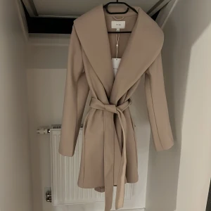 Beige kappa med bälte från Vila - Stilren beige kappa från Vila med bred krage och knytskärp i midjan. Jackan har lång ärm och en klassisk, rak passform som ger en snygg siluett. Perfekt för höst och vinter! Aldrig använd med prislapp kvar! 