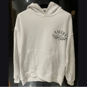 Vit hoodie från Amiri - Säljer en stilren vit hoodie från Amiri med tryck både fram och bak. Hoodien har en klassisk design med känguruficka och långa ärmar. Perfekt för en avslappnad look.