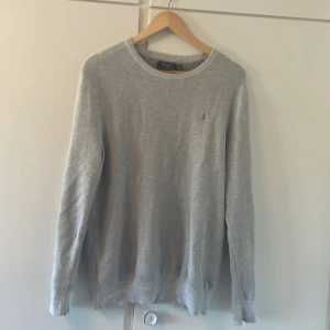 Grå stickad tröja från Polo Ralph Lauren - Snygg grå stickad tröja från Polo Ralph Lauren med klassisk rund hals och diskret broderad logga på bröstet. Långärmad modell i mjuk bomull som passar perfekt till jeans eller chinos. Enkel och stilren vibe för dig som gillar clean look.