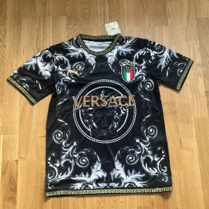 Italien Versace x Puma fotbollströja - Unik svart fotbollströja från Puma i samarbete med Versace och Italien. Tröjan har korta ärmar, guldiga detaljer, stort Versace-tryck och Medusa-motiv på bröstet. Mönstret är barockinspirerat i vitt och grått, med italienska landslagets emblem och klassiska Versace-bårder.