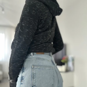 Svart hoodie från Champion med snörning - Svart hoodie från Champion med vita spräckliga detaljer och vit snörning i huvan. Tröjan har en klassisk magficka och tryckt logga framtill. Perfekt för dig som gillar en sportig och avslappnad stil.