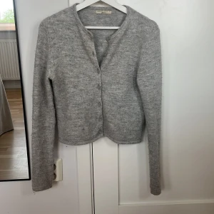 Grå stickad kofta från Gina Tricot - Mysig grå stickad kofta från Gina Tricot i storlek XS. Koftan har rund halsringning, långa ärmar och knäppning framtill med matchande knappar. Perfekt för lager-på-lager och enkel att styla till olika outfits.