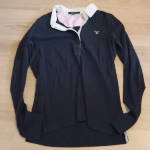 Mörkblå pikétröja från Gant - Snygg mörkblå långärmad pikétröja från Gant med vit krage och broderad logga på bröstet. Tröjan har klassisk pikélook med knappslå och insida i ljusrosa/vit randigt tyg. Perfekt för en preppy stil.