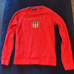 Röd långärmad tröja från GANT - Röd långärmad tröja från GANT med broderad logga och sköld på bröstet. Klassisk rund halsringning och mjukt bomullsmaterial. Perfekt för dig som gillar sportig och stilren look. Passformen är normal och tröjan har ribbade muddar vid ärmslut och nederkant.