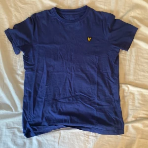 Blå t-shirt från Lyle & Scott - Snygg marinblå t-shirt från Lyle & Scott med klassisk gul logga på bröstet. T-shirten är i mjuk bomull och har rund hals och korta ärmar. Perfekt basic-plagg med stilren look och skön passform.