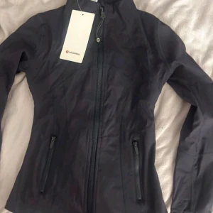 Svart define jacket från Lululemon - Snygg svart vindjacka från Lululemon med figurnära passform. Jackan har hel dragkedja framtill, två dragkedjeförsedda fickor och hög krage. Perfekt för dig som gillar sportig och clean stil. Materialet är lätt och smidigt, perfekt för aktiva dagar.