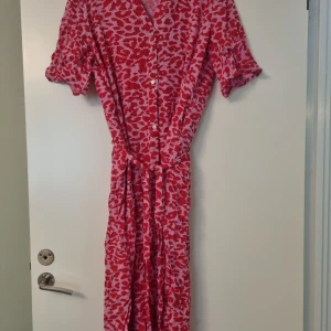Rosa och röd mönstrad klänning Saint - Snygg långklänning från Saint i rosa och rött med leopardmönster. Klänningen har korta ärmar, knappar framtill, v-ringning och ett matchande bälte i midjan. Perfekt för dig som vill sticka ut med färg och mönster.