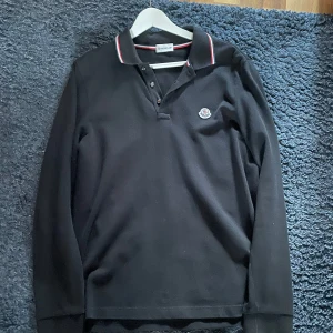 Svart Moncler pikétröja långärmad - Svart långärmad pikétröja från Moncler med klassisk krage och knäppning framtill. Kragen har snygga röda och vita ränder och Moncler-loggan sitter på bröstet. Storlek M