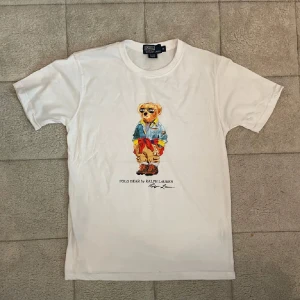 Vit Polo Bear t-shirt från Ralph Lauren - Vit t-shirt från Polo Ralph Lauren med ikoniskt Polo Bear-tryck på bröstet. Klassisk rund hals och korta ärmar. Tillverkad i mjuk bomull som känns skön mot huden. Perfekt för dig som gillar streetstyle och vill sticka ut med en cool print.